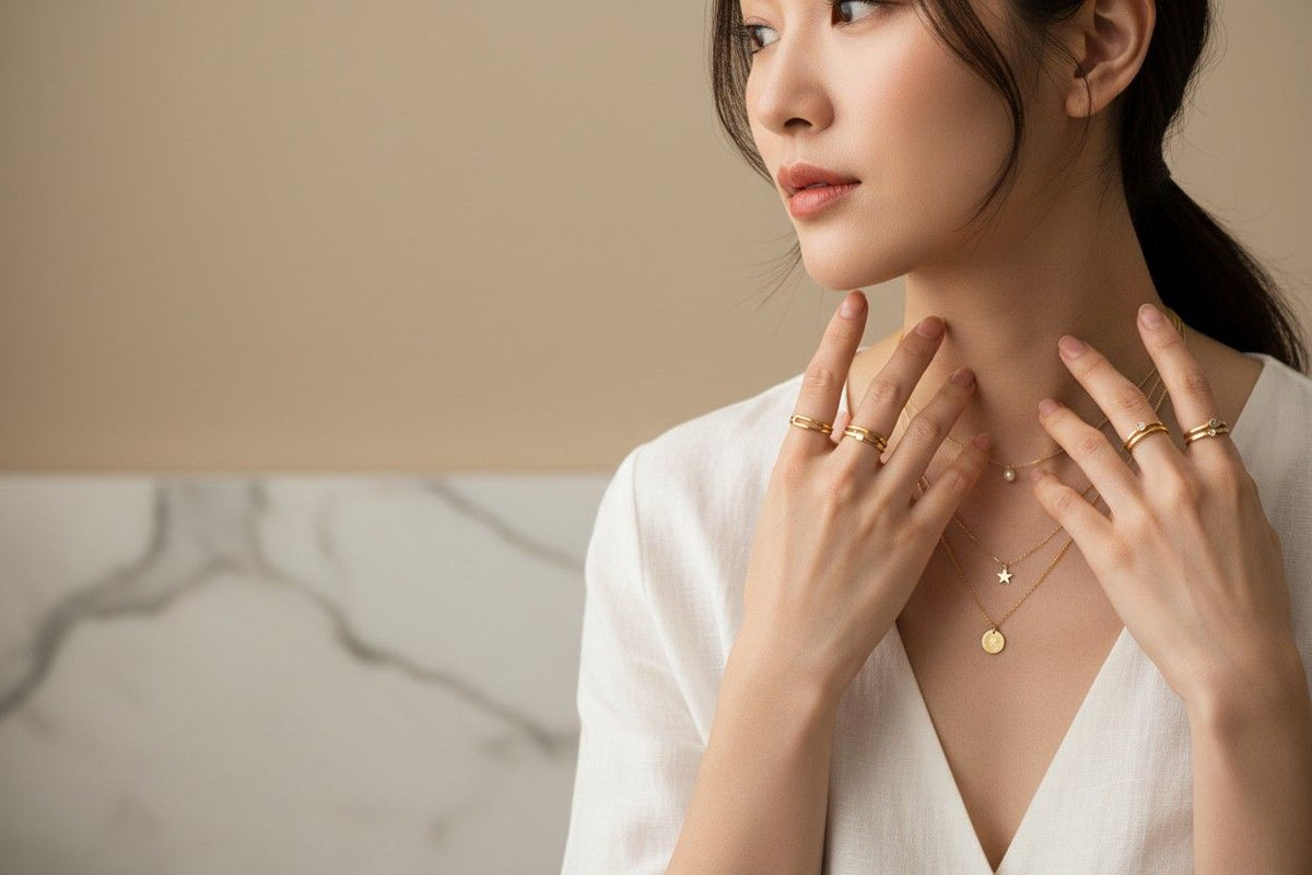 Woman wearing gold jewelry, including a necklace and rings, against a neutral background.